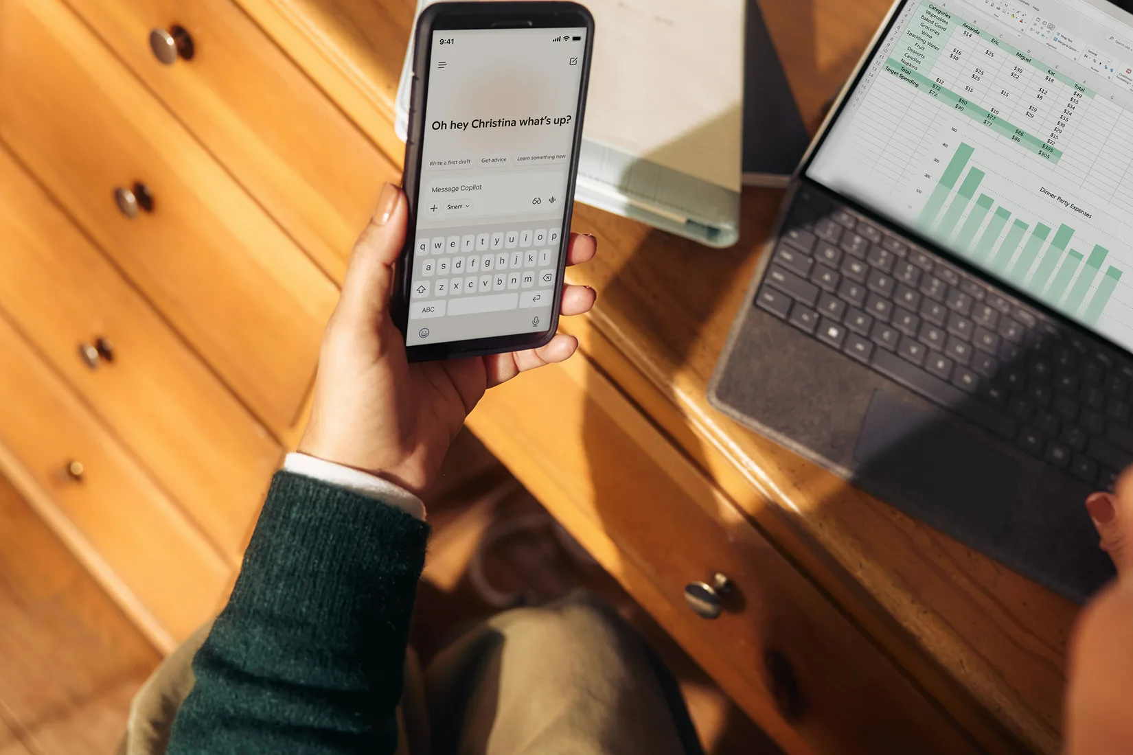 Overhead view of a hand holding a smartphone with an AI chat app beside a laptop showing a budget spreadsheet on a wooden desk